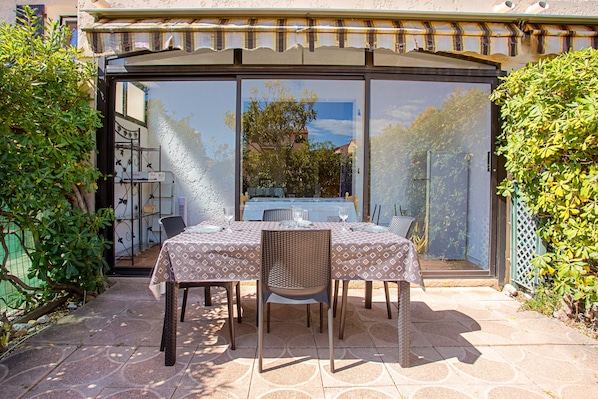 An outdoor patio with a table set for two, surrounded by chairs, and a glass door leading to a garden.