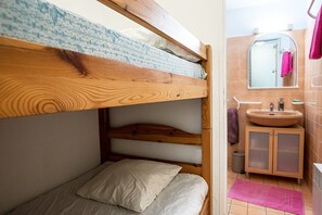 Ein Etagenbett mit einem gemusterten Kopfteil, ein kleiner Nachttisch, ein Badezimmer mit Waschbecken und Spiegel sowie ein pinkfarbener Teppich auf dem Boden.