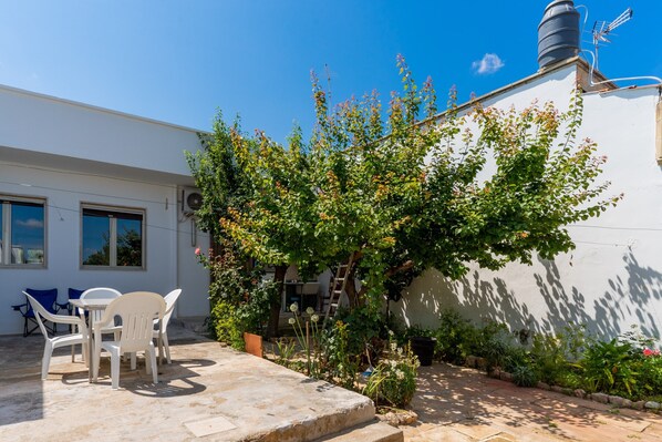 Un patio con mobiliario exterior, un edificio blanco y un jardín con diversas plantas y un árbol.