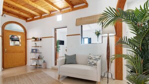 A living room with a sofa, a wooden ceiling, a door, a shelf with items, and a plant.