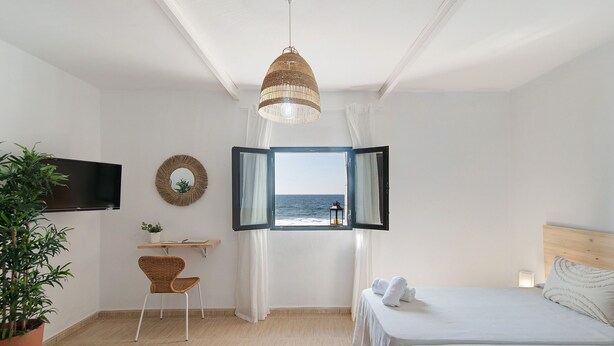 A bedroom with a bed, a desk, a chair, a TV, a window with curtains, and a view of the sea.