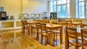 Un comedor con mesas y sillas de madera, una barra de buffet con alimentos, y ventanas amplias que dejan entrar luz natural.