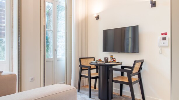 Una habitación de hotel moderna con un ventanal grande, un televisor de pantalla plana montado en la pared, una mesa redonda con dos sillas y una mesita con un frutero.
