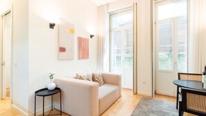 Una sala moderna con un sofá beige, una mesita auxiliar negra y un jarrón con flores. Hay dos cuadros abstractos en la pared y una zona de comedor con una mesa y sillas.