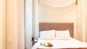 Una cama bien tendida con sábanas y almohadas blancas, una mesita de noche con una taza y un libro, y una bandeja con croissants y fruta.