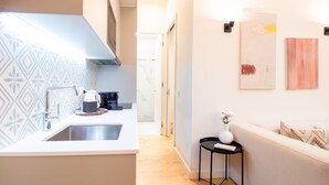 Una cocina moderna con horno empotrado, cafetera y lavamanos. Un pasillo conduce a una sala de estar con sofá y una mesita.