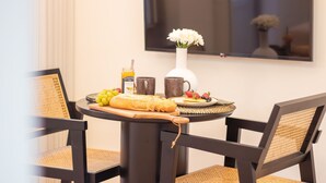 Una mesa redonda para comer con una hogaza de pan, una botella de miel y un plato de fruta. Dos sillas con asientos de mimbre están colocadas alrededor de la mesa.