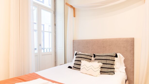 Una cama bien hecha con un cabecero color beige, dos almohadas estampadas y una toalla blanca doblada sobre una manta naranja.