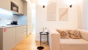 Cocina moderna con microondas, hervidor y cafetera. Hay una pequeña mesa redonda con un florero y una taza. Un sofá color beige con cojines estampados se encuentra en la zona de estar.