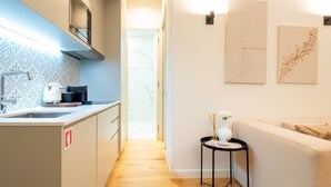 Una cocina moderna con horno empotrado, cafetera y un lavabo. Hay una pequeña mesa redonda con un florero y una taza. El espacio tiene una paleta de colores neutros, con un sofá beige y una pared blanca.