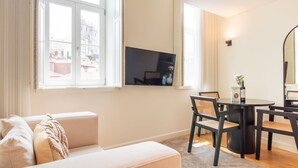 Moderni olohuone, jossa on sohva, ruokapöytä tuoleineen, seinälle asennettu taulutelevisio ja suuret ikkunat, joista avautuu näkymä kaupunkimaisemaan.
