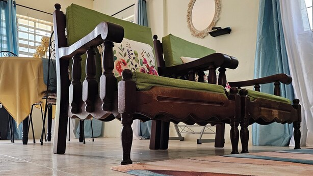 Una habitación con un sofá de madera, un cojín verde y una almohada estampada. Hay una mesa con mantel amarillo y una silla. El piso es de baldosas y la ventana tiene cortinas.
