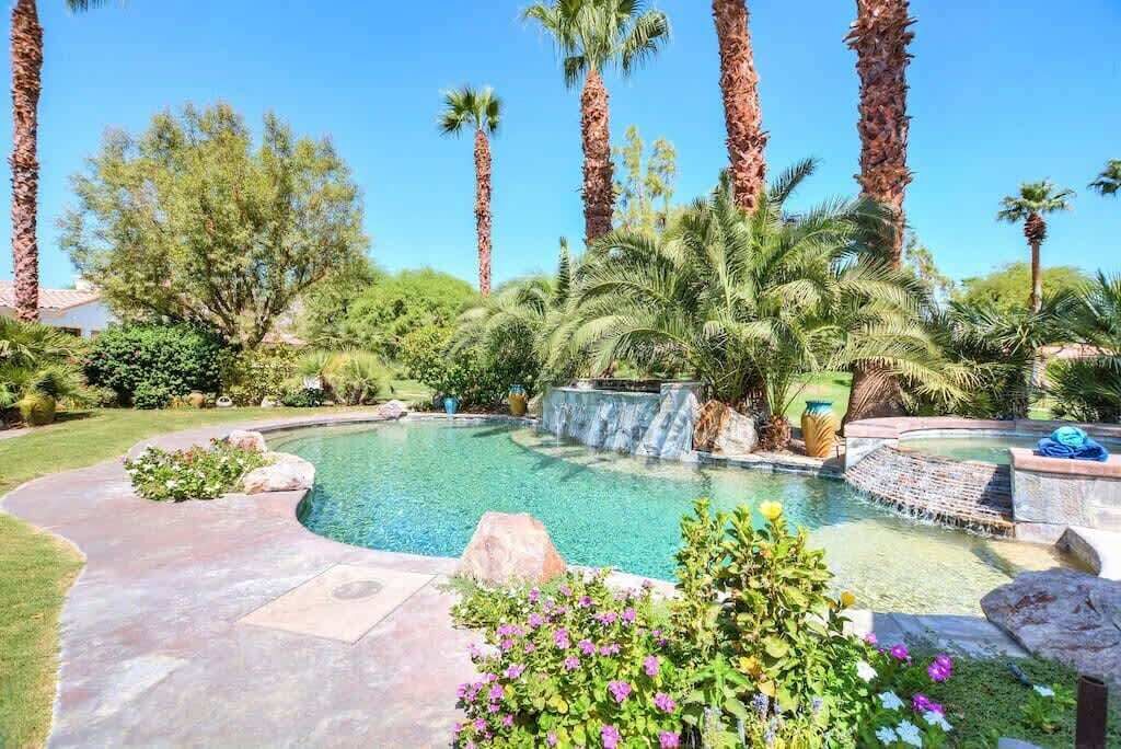 A swimming pool surrounded by palm trees and greenery, with a waterfall feature and a stone pathway.