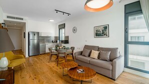 Un salon moderne avec un canapé, une table basse et une table à manger. On aperçoit au fond une cuisine équipée d’un réfrigérateur et d’une fenêtre.