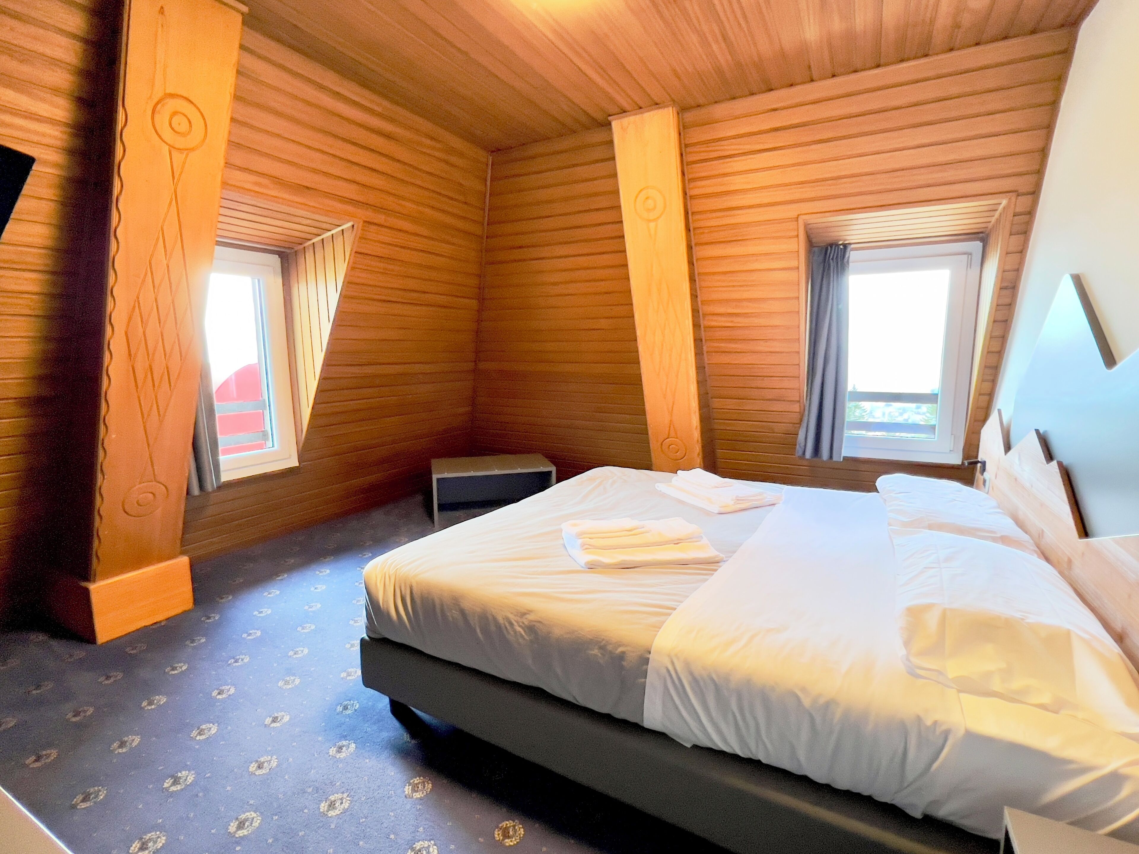 A wooden room with a bed, two windows, and a small table.