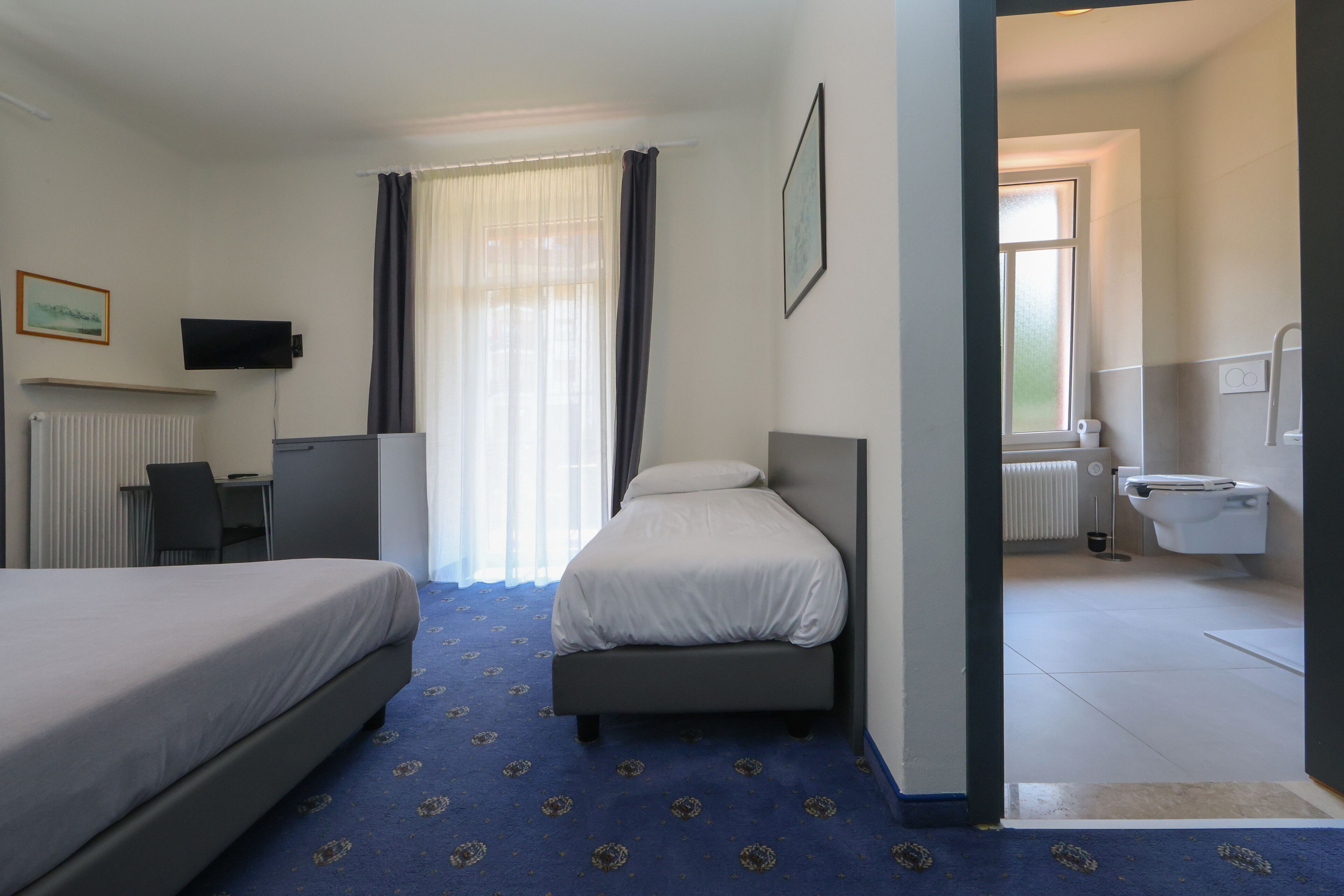 Ein Hotelzimmer mit zwei Betten, einem Schreibtisch, einem Fernseher, einem Fenster mit Vorhängen und einem Badezimmer mit Toilette und Heizkörper.