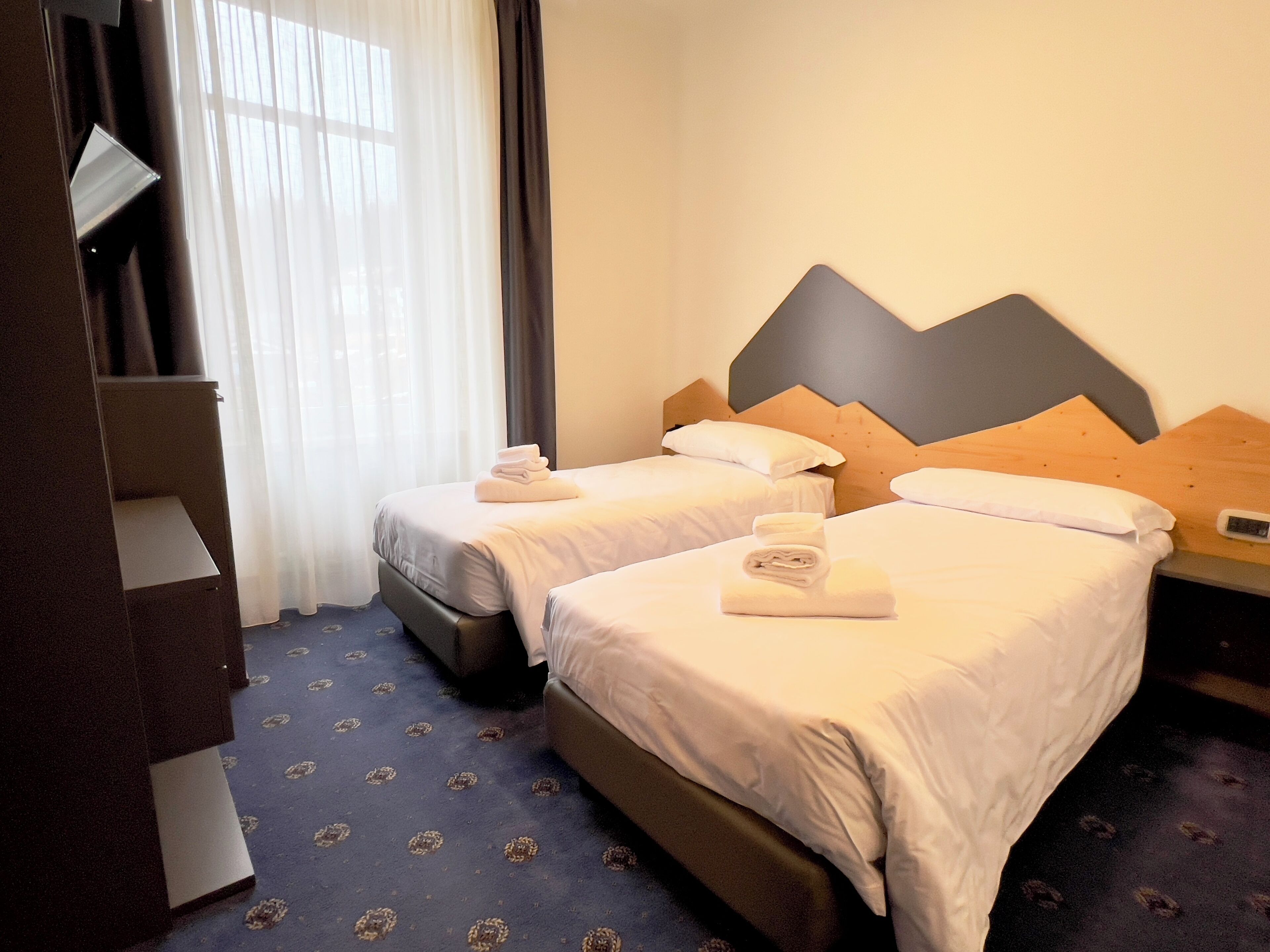 A hotel room with two beds, a wooden headboard, a desk, and a window with curtains.
