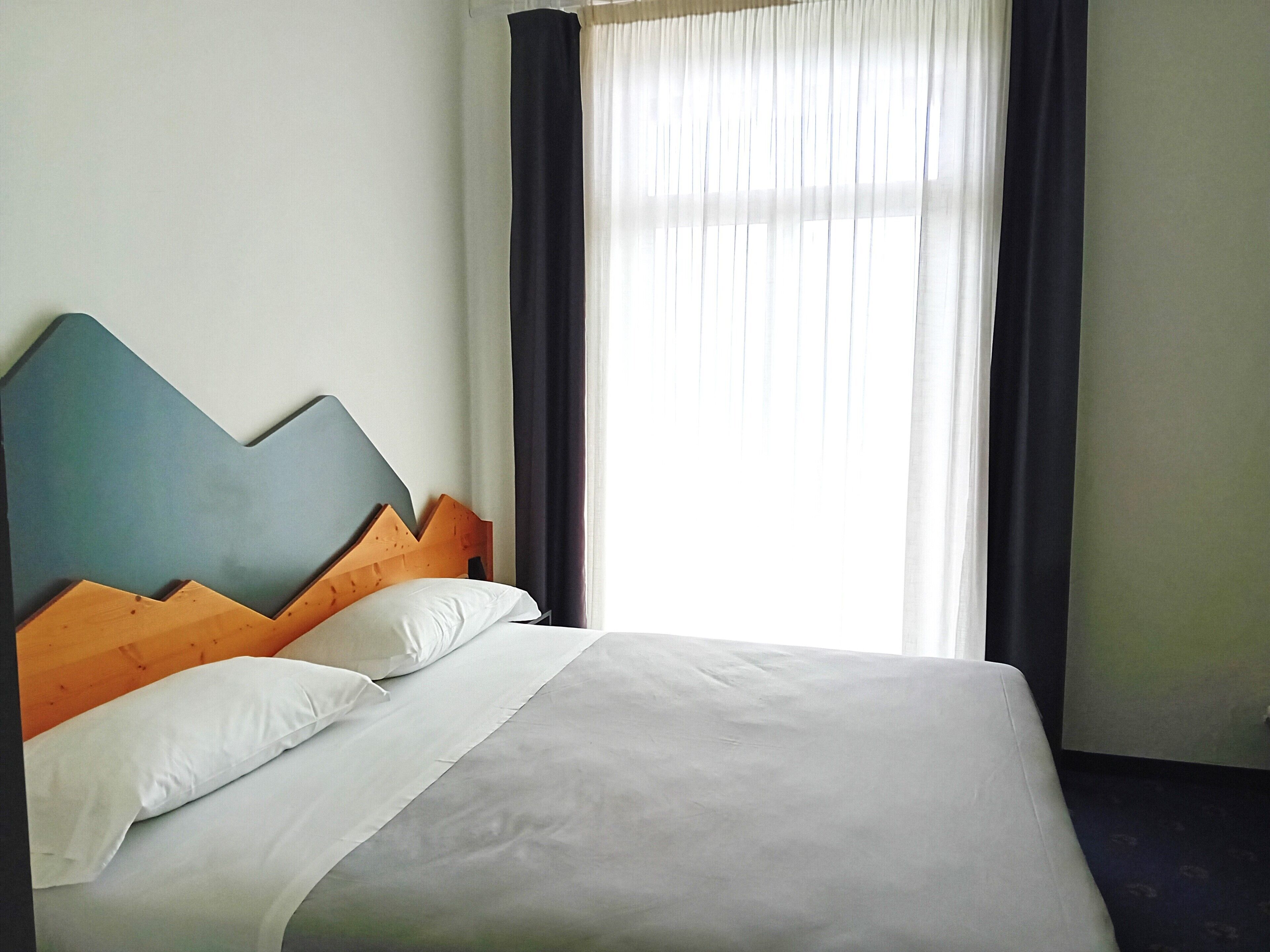 Una camera d'albergo con un letto, una testiera in legno, biancheria da letto bianca e una finestra con tende.