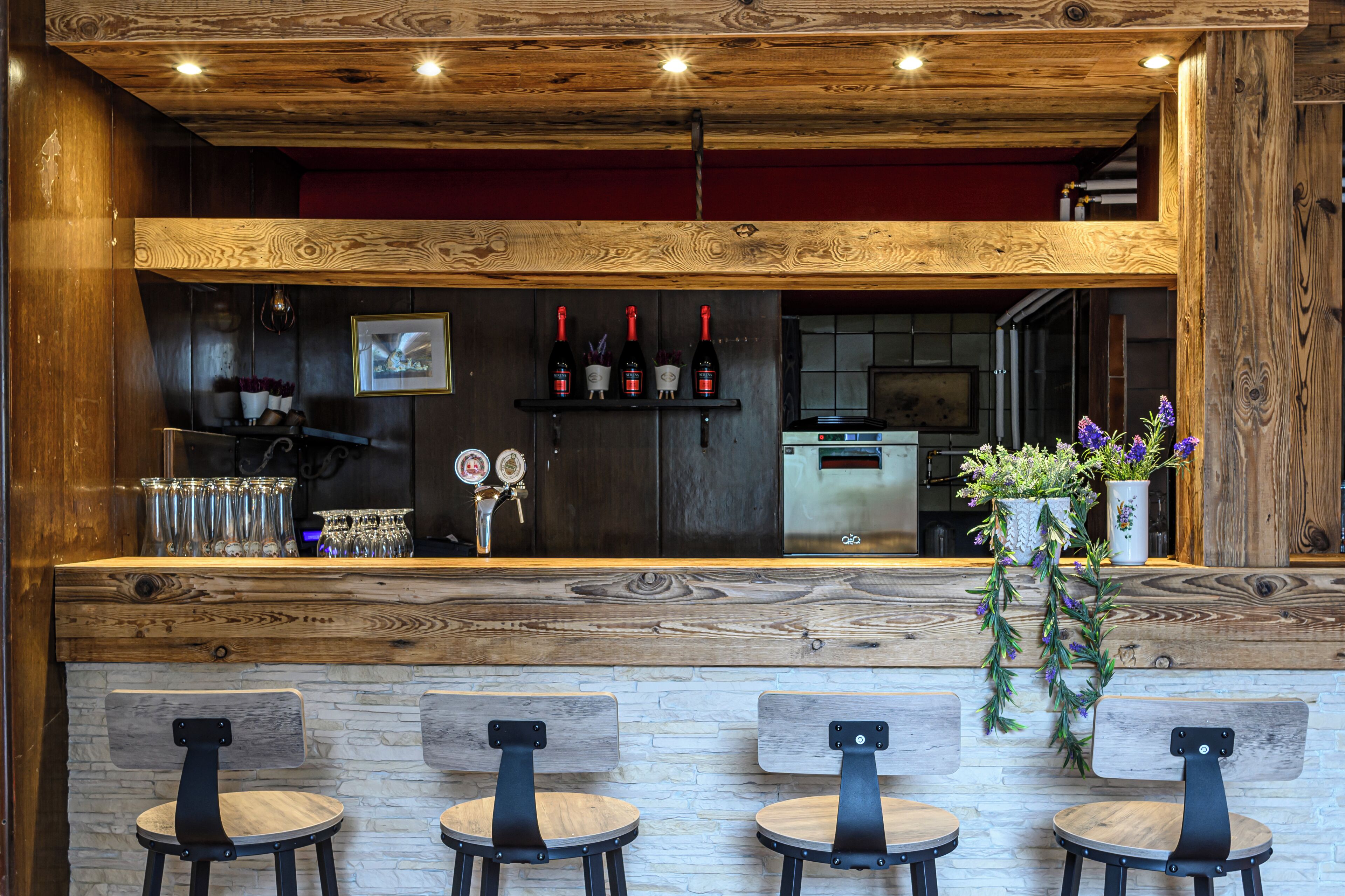 Zona de bar con techo de madera, una barra con botellas, vasos y un refrigerador. Hay cuatro taburetes y una planta en maceta con flores.