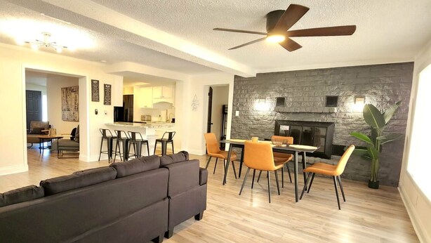 Ein modernes Wohnzimmer mit einem dunkelgrauen Sofa, einem Essbereich mit orangefarbenen Stühlen, einer Wand aus Naturstein und einem Deckenventilator.