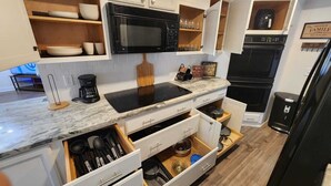 Eine moderne Küche mit einer schwarzen Mikrowelle, einem eingebauten Ofen und einer schwarzen Induktionskochplatte. Weiße Schränke mit Holzregalen befinden sich ober- und unterhalb der Arbeitsplatte.