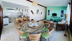 A spacious living area with a round glass table surrounded by wicker chairs, a kitchen with white cabinets, and a teal accent wall. - Complete house in Maresias, 450m from the sand (SÃO SEBASTIÃO)