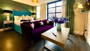 Ein Hotelzimmer mit einem Bett, einem violetten Sofa, einem kleinen Tisch mit Blumenvase und einem Fenster mit Vorhängen.