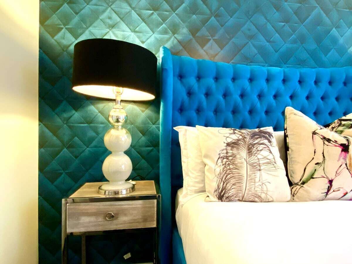Una cama con cabecera capitonada, una lámpara de noche y almohadones decorativos.