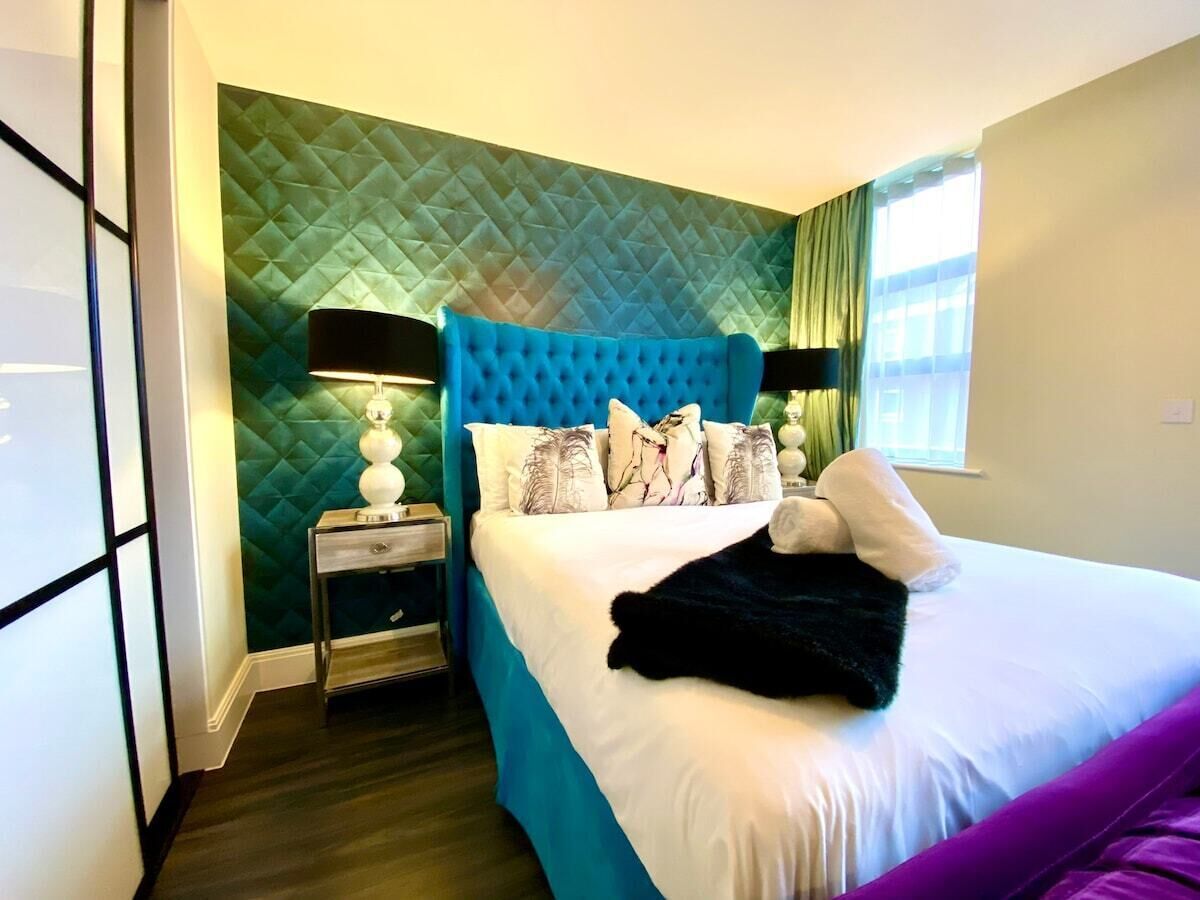 Een moderne hotelkamer met een gestoffeerd hoofdeinde, een groot bed met meerdere kussens, een nachtkastje met een lamp en een groen, textuurrijk wand.