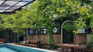 A swimming pool with tiled edges, surrounded by wooden benches and picnic tables, with a brick wall and glass panels in the background. - The Wishes Hotel At Chiangmai Chiang Mai (Chiang Mai)
