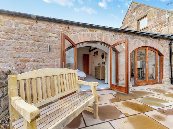A wooden bench in a stone building with an open archway leading to a room with a bed and dresser.