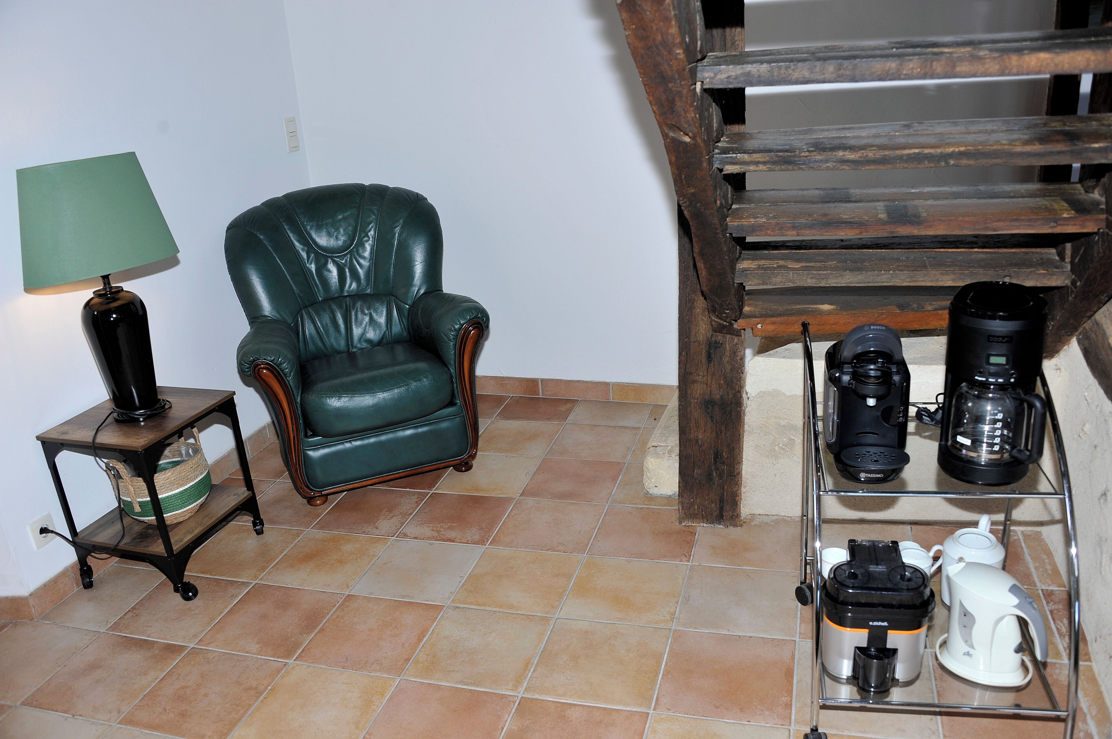 Een kamer met een groene leren fauteuil, een tafeltje met een lamp en een plank met koffiezetapparaten en een waterkoker.