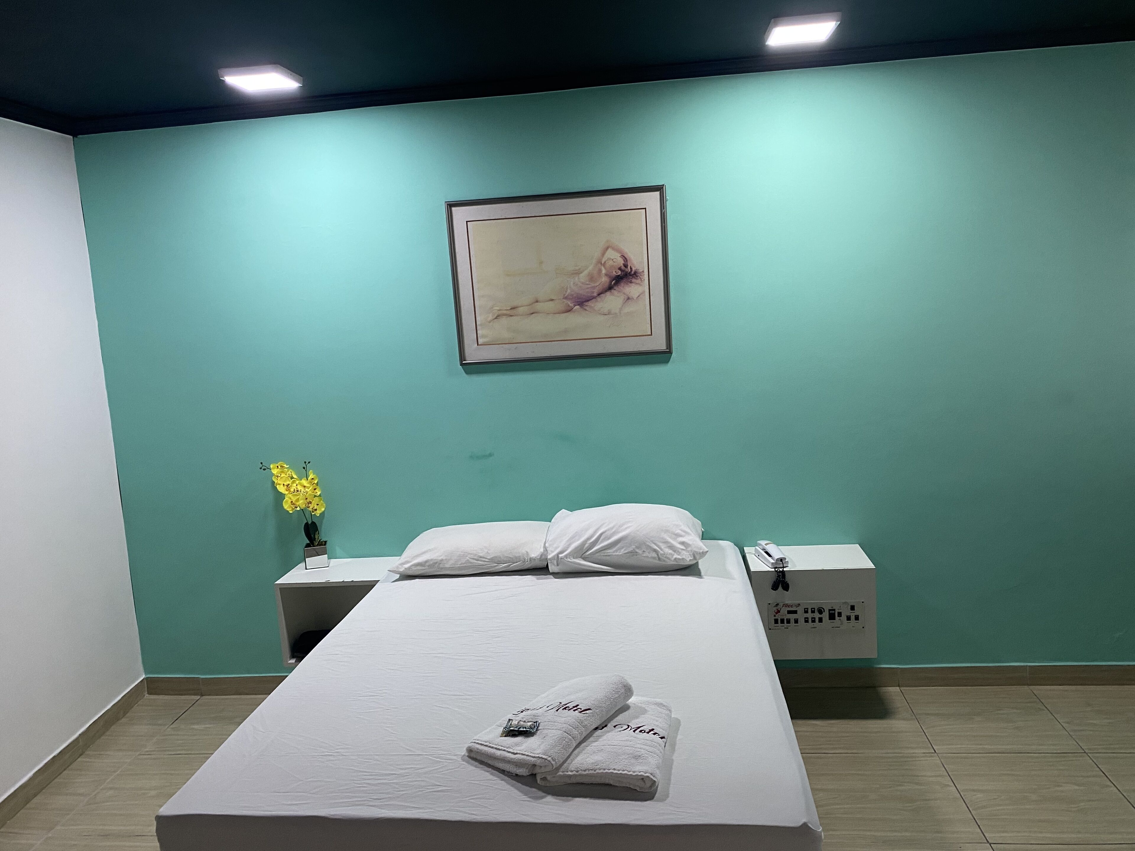 A hotel room with a bed, a nightstand, a vase with flowers, and a painting on the wall.