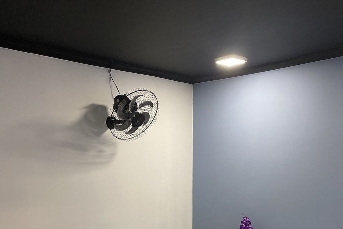 A room with a bed, a desk, two chairs, a nightstand with a lamp, and a wall-mounted fan.