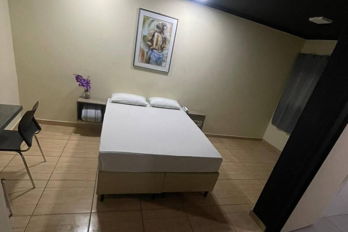 A bedroom with a bed, a desk, a chair, a small shelf with a vase, and a painting on the wall.