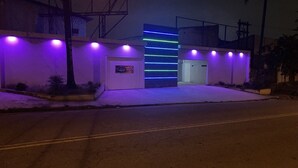 A building with a modern facade, illuminated by purple and blue lights, featuring a central entrance and a small garden area. - Light Motel (Guarulhos)