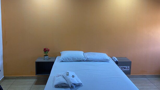 A hotel room with a bed, white pillows, a bedside table with a phone, a small vase with flowers, and a power strip. - Light Motel (Guarulhos)