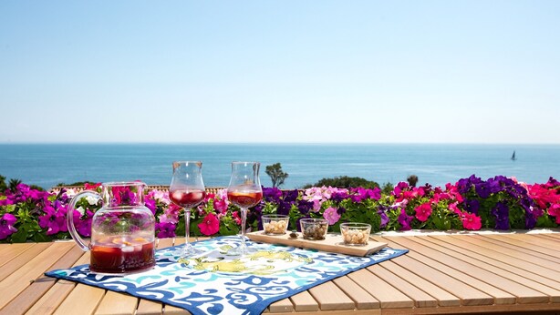 Et bord med en kande og glas med rød drik, en bakke med snacks, og udsigt til havet.