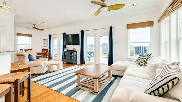 A spacious living room with a large sofa, a coffee table, and a TV. There are ceiling fans, a dining area with chairs, and a view through large windows. - Bluefin - Amazing Panoramic Sea, Bridge, Bay Views! (Freeport)
