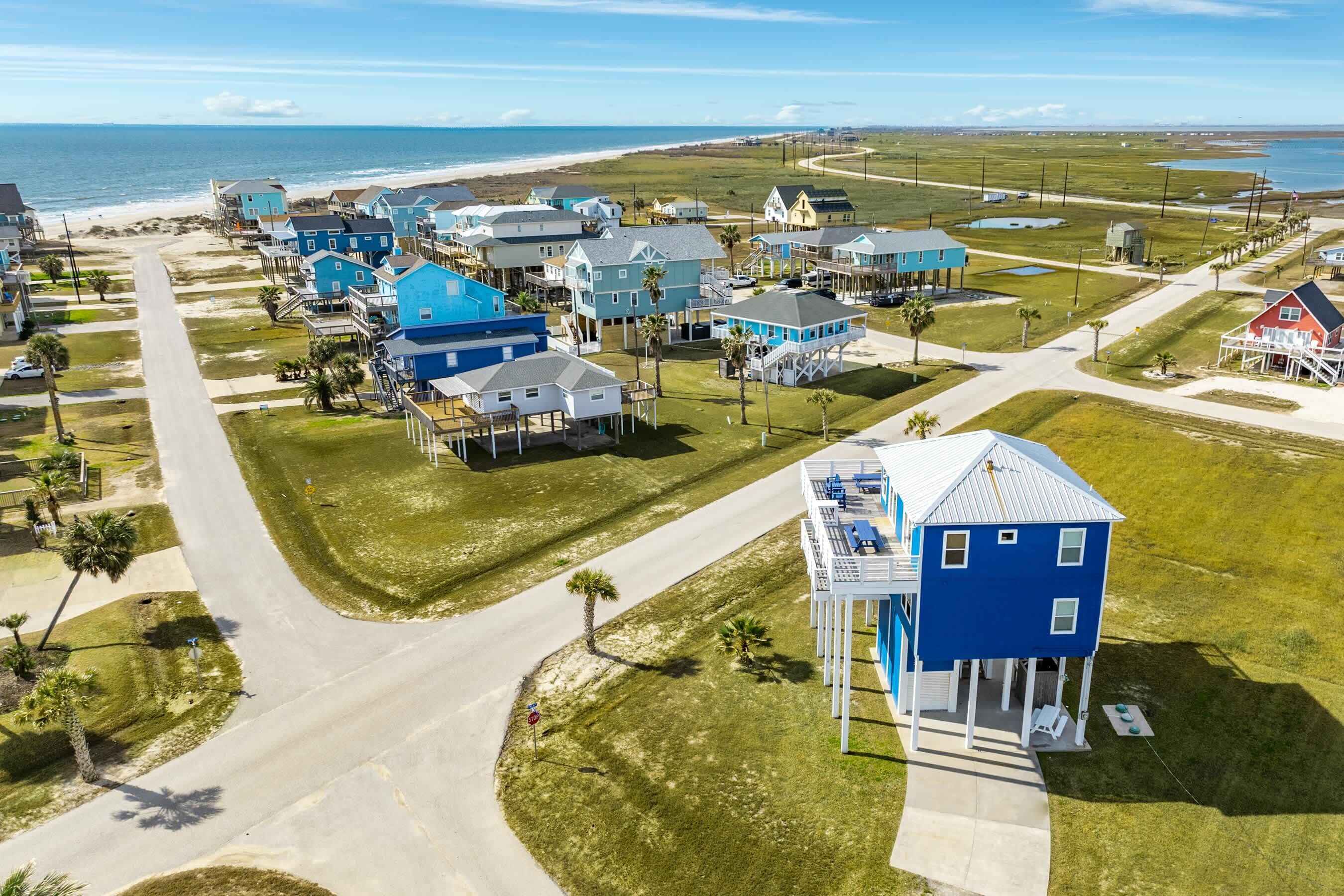A coastal residential area with houses featuring blue exteriors, a beach in the background, and a clear sky.