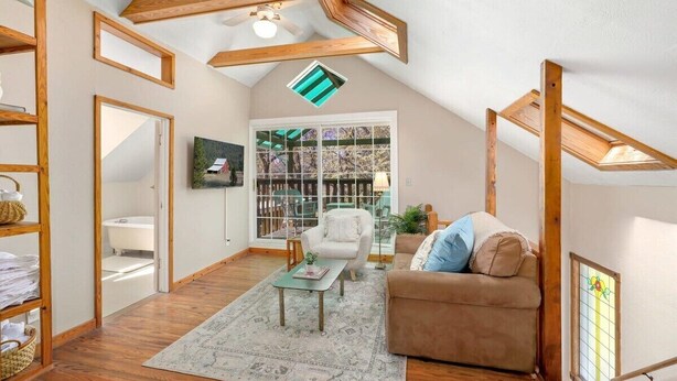 A cozy attic room with wooden beams, a ceiling fan, a large window with a view of a deck, a brown sofa, and a small table with a rug. - Cottage on Rush - Heart of Fayetteville (Fayetteville)
