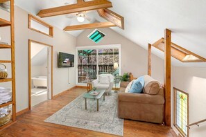 Un acogedor dormitorio en ático con vigas de madera, ventilador de techo, un ventanal con vistas a una terraza, un sofá marrón y una pequeña mesa con alfombra.
