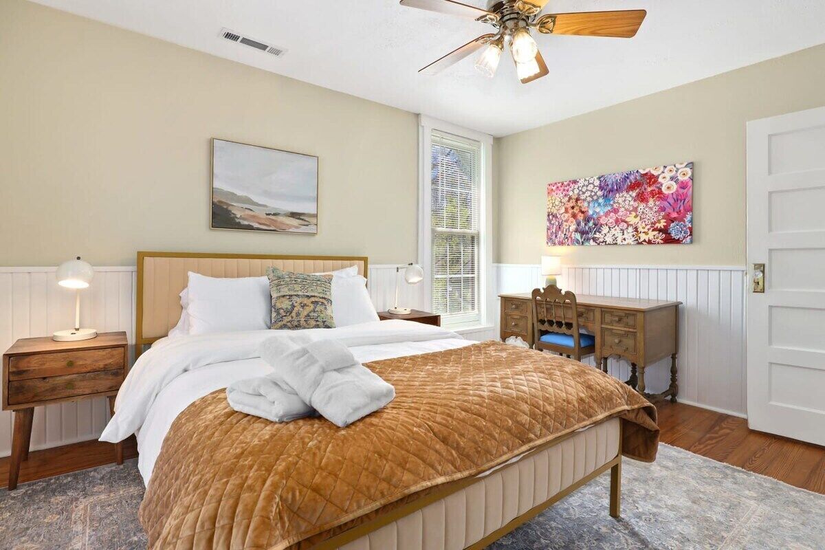 A bedroom with a bed, bedside table, nightstand, desk, chair, and a ceiling fan.