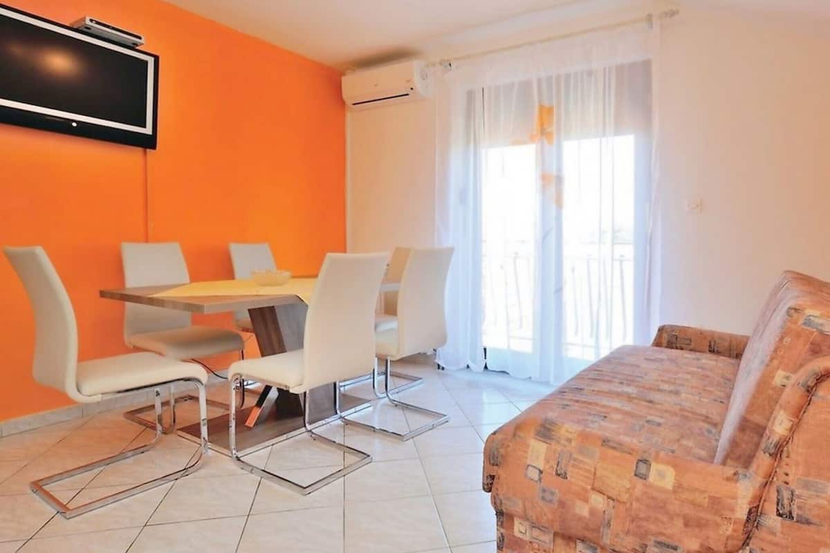 A modern living room with a sofa, a dining table with chairs, a flat-screen TV mounted on the wall, and a large window with curtains.