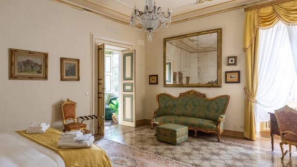 A room with a bed, a sofa, a chair, a mirror, and a chandelier.