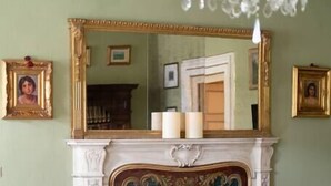 A room with a fireplace, floral-patterned chairs, a chandelier, and framed pictures on the walls.