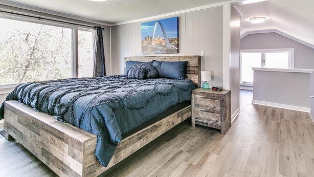 Une chambre à coucher avec un grand lit, une tête de lit en bois, des tables de chevet et une vue sur le paysage urbain par la fenêtre.