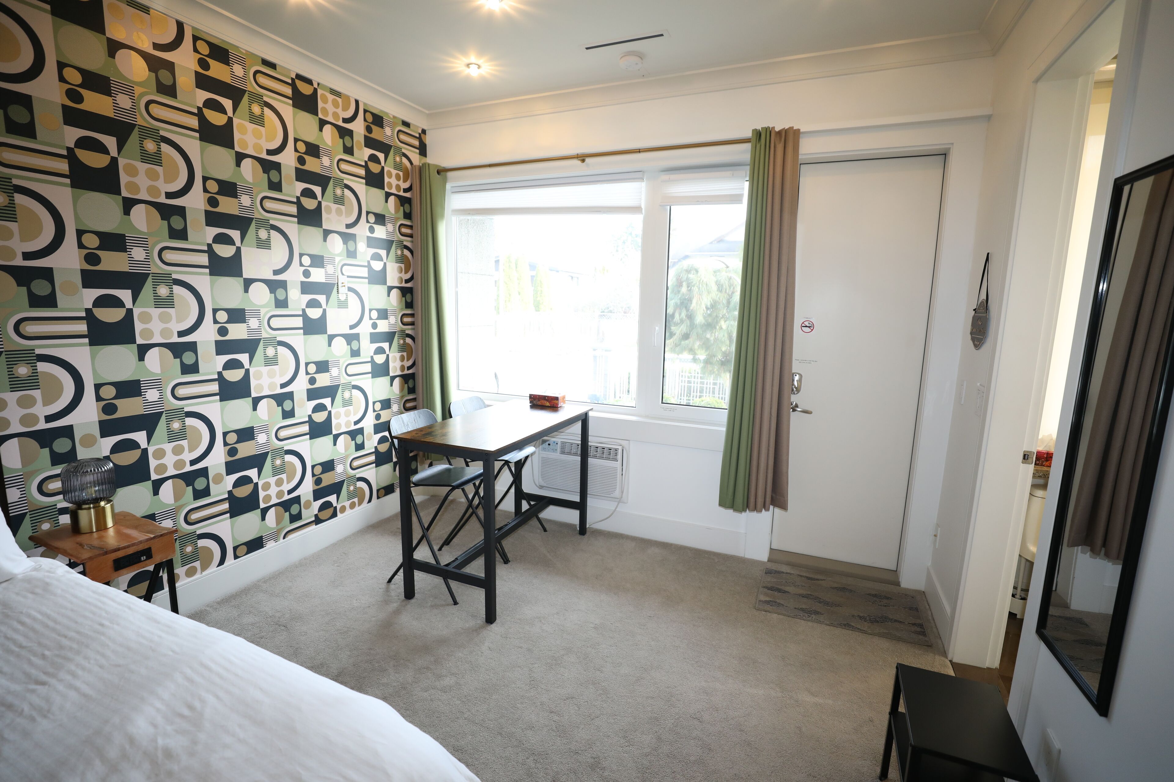 A room with a patterned wallpaper, a bed, a desk, a chair, a window with curtains, and a door.