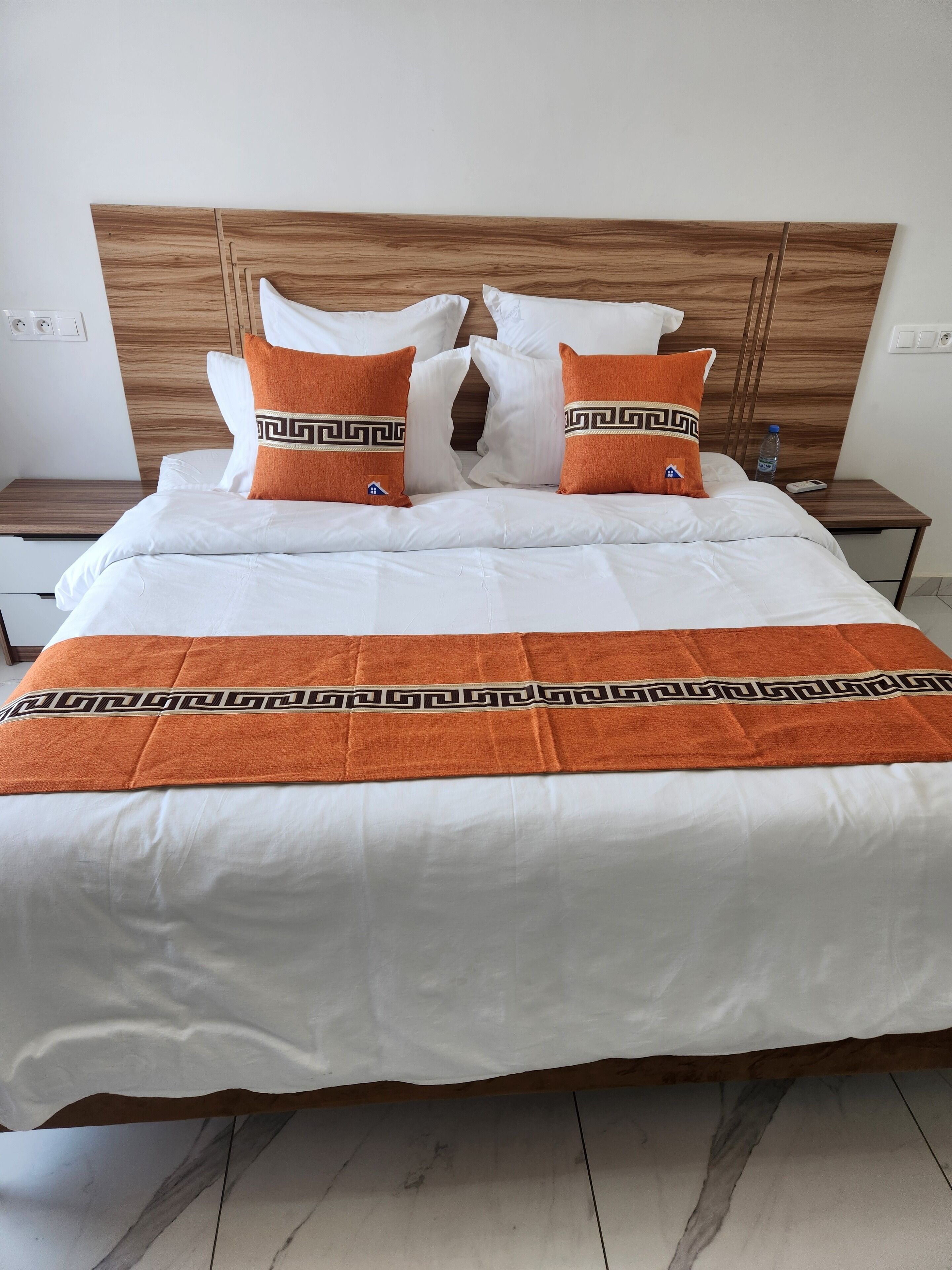 Un lit bien fait, avec du linge de lit blanc et deux coussins orange ornés d’un motif géométrique.