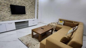 A modern living room with a sectional sofa, a coffee table, a flat-screen TV mounted on a textured wall, and a minimalist white cabinet. - MARINA APPART HÔTEL  (Dakar)